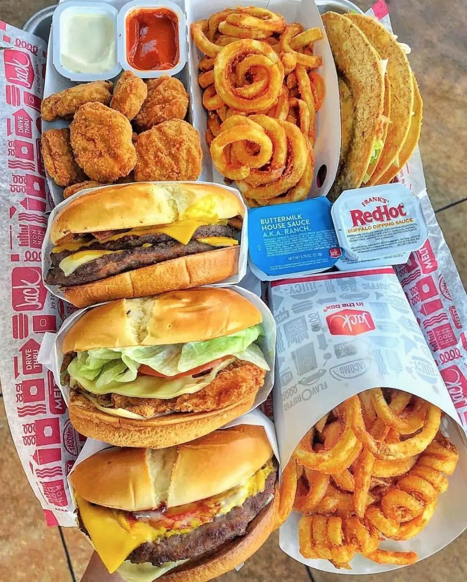 Overhead view of fast food meal spread including fried chicken, burgers with cheese, curly fries, onion rings, tacos, and dipping sauces on branded packaging
