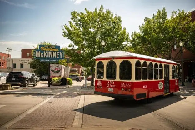 Red and white trolley driving in front of a neon sign that says Welcome to Historic Downtown McKinney
