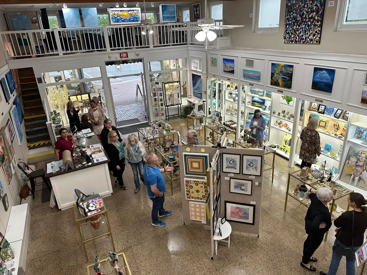 Interior of art gallery with visitors viewing framed artwork and paintings displayed on walls and easels throughout the two-story space