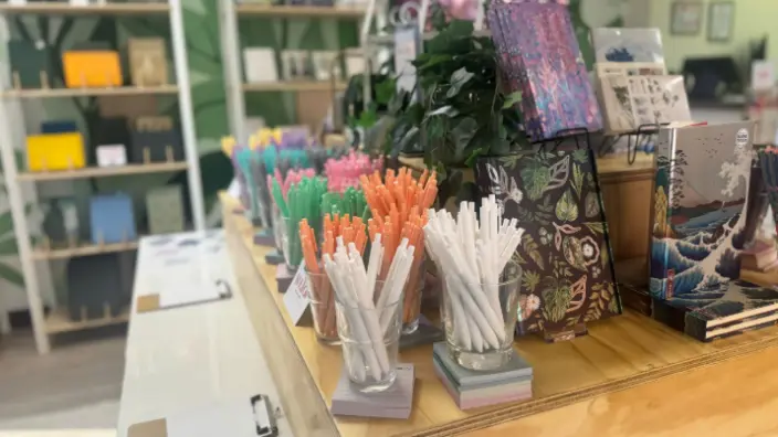 Colorful pens and markers displayed in containers on a wooden counter, with shelves of books and supplies visible in the background
