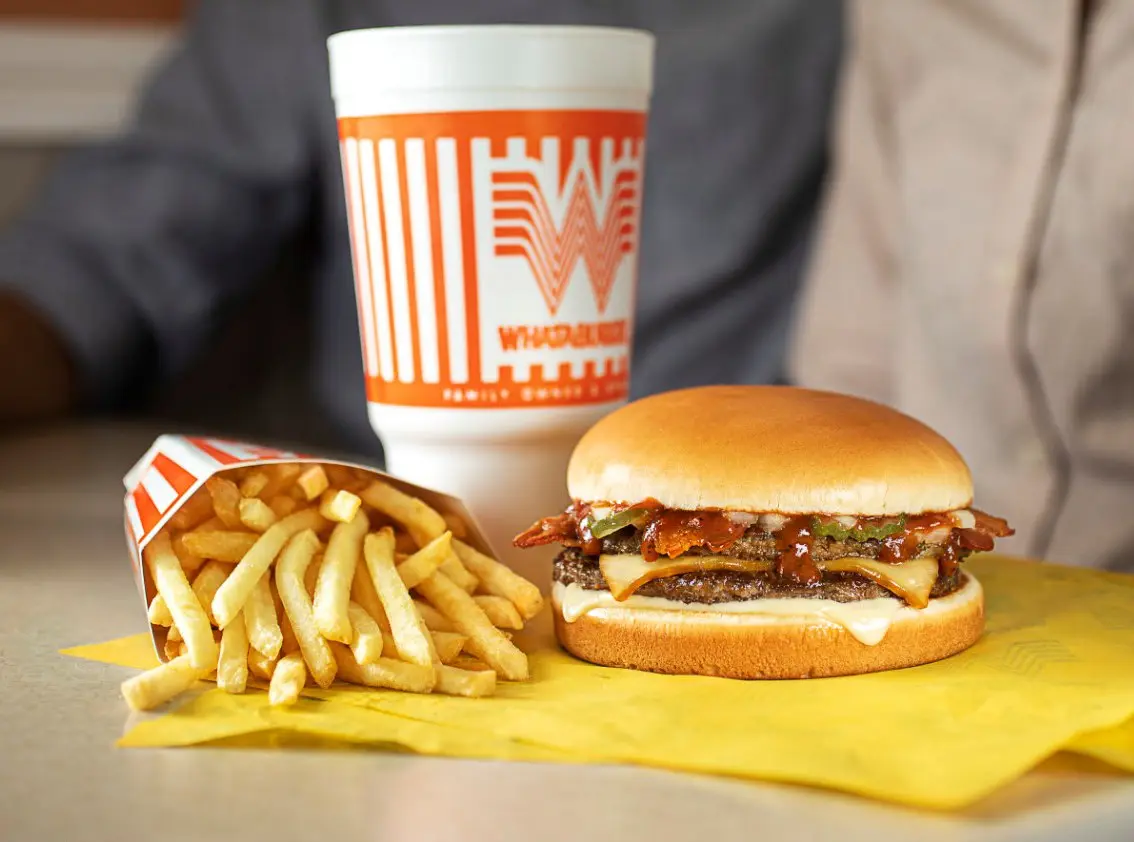 Cheeseburger with crispy fries and branded drink cup on yellow wrapper, held by person in dark clothing
