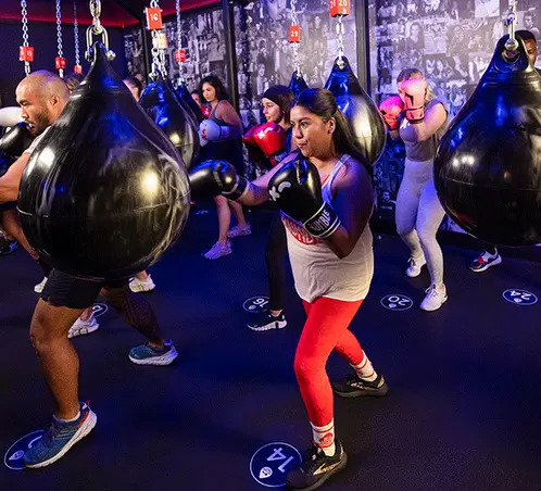 Group fitness class participants punching large suspended heavy bags under blue lighting in a boxing studio