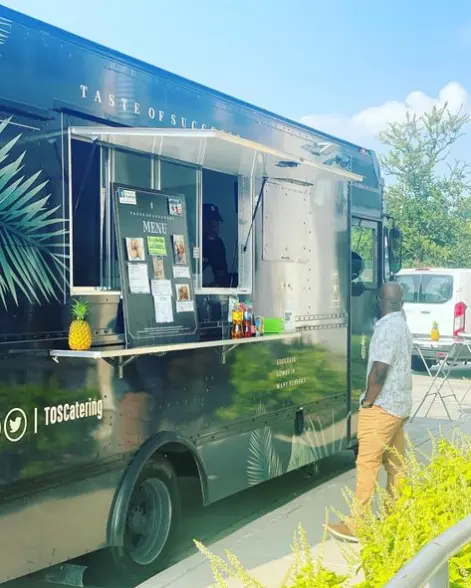 Dark green food truck with service window, person standing at counter, pineapple decoration visible, outdoor setting with trees and grass