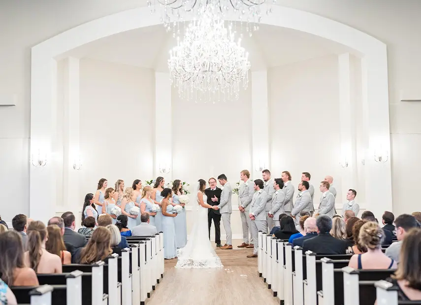 Wedding ceremony in white chapel with arched ceiling, crystal chandelier, guests seated in pews facing bridal party at altar