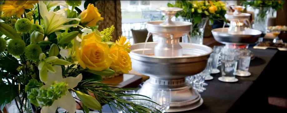Yellow and white flowers in a vase on a table with silver serving bowls and glassware in a dining venue
