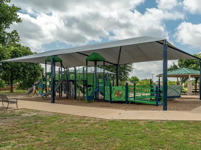 Green and blue playground structure with shade canopy in open park setting surrounded by grass and trees