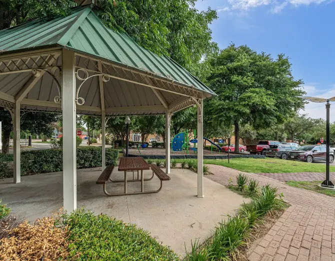Covered picnic pavilion with green metal roof and attached picnic table in park setting surrounded by trees and parked cars