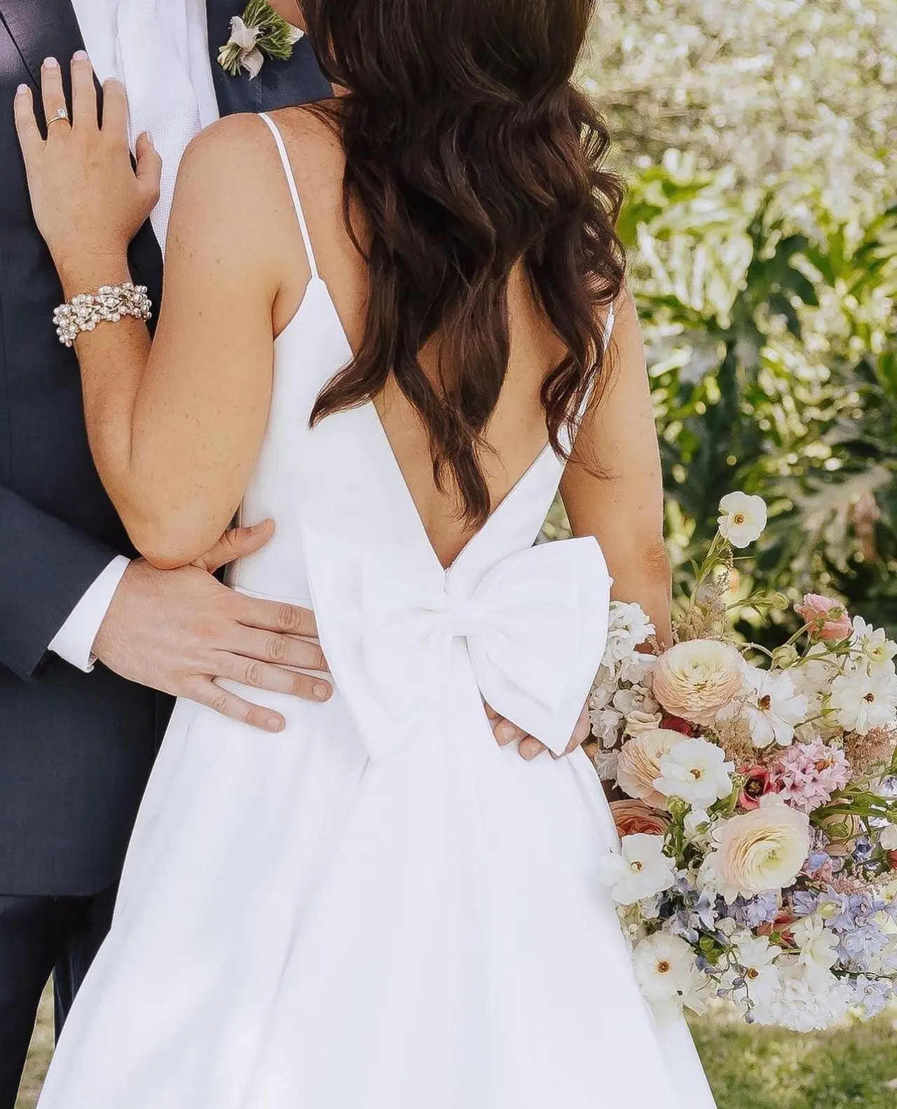 Bride in white gown with bow detail and beaded bracelet embraced by groom, holding bouquet of cream and pink flowers