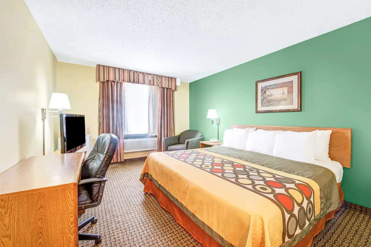 Hotel bedroom with green accent wall, queen bed with patterned comforter, desk with TV, and window with curtains
