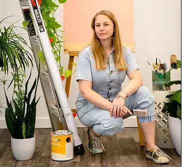 Woman with long reddish-blonde hair sitting on wooden stool in art studio surrounded by potted plants and art supplies