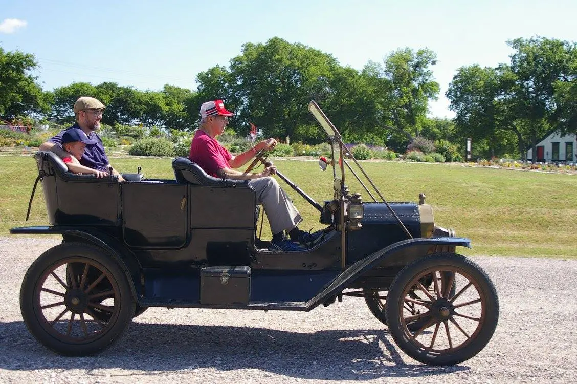 Three people riding in an open-air vintage automobile on a paved path, with green fields and trees in the background