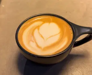 Latte with heart-shaped latte art in a black mug on tan surface