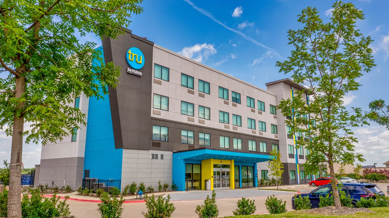 Modern hotel building with blue and tan facade, ground-level entrance, and surrounding landscaping under clear sky