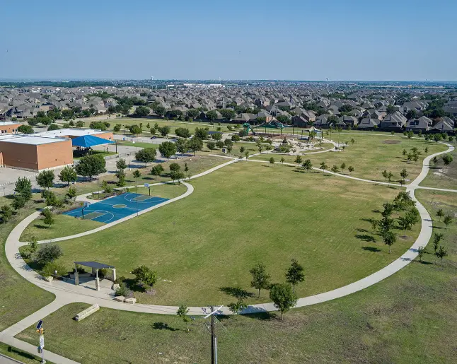 Aerial view of expansive park with blue swimming pool, brick building, green lawn, curved pathways, and residential neighborhood in distance