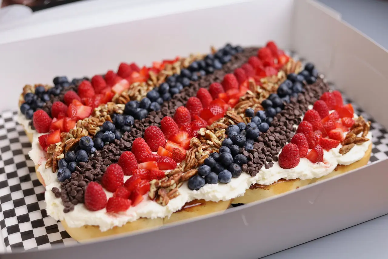 Rectangular pastry topped with whipped cream, red raspberries, dark blueberries, and granola stripes, served on checkered parchment