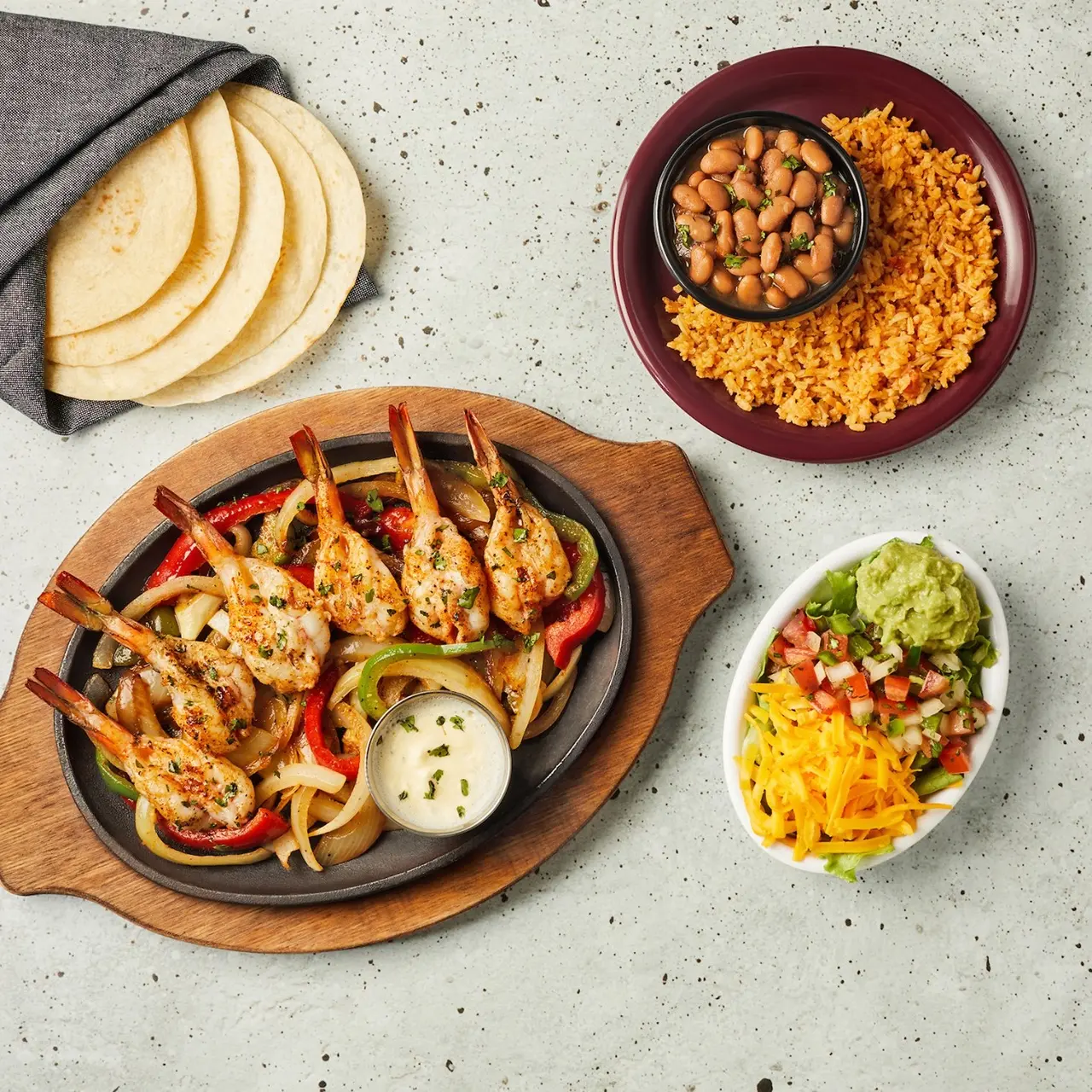 Sizzling shrimp fajitas on cast iron with peppers, sliced apples, Mexican rice, chickpeas, and fresh salsa on concrete surface