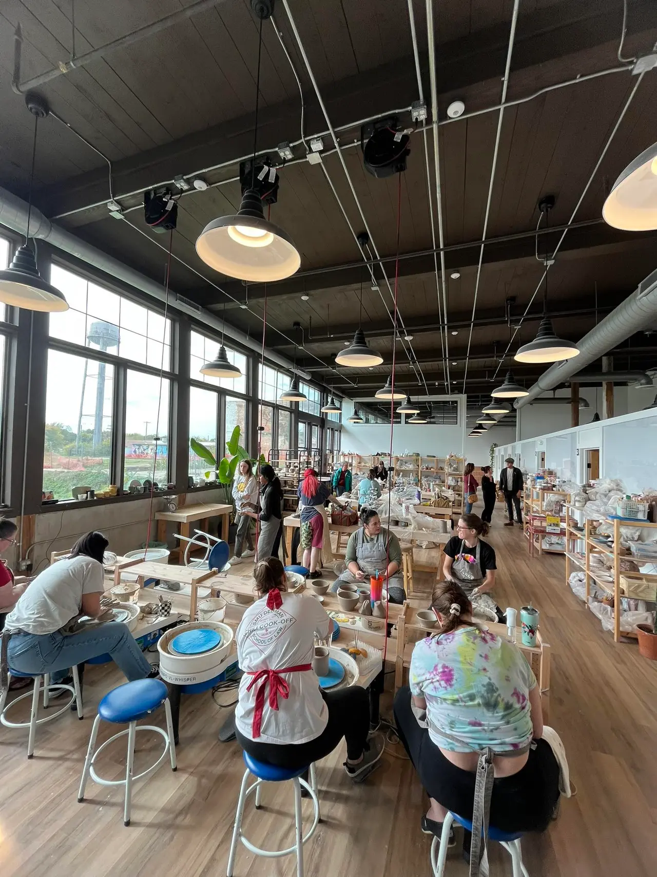 Spacious ceramic studio with high ceilings and large windows, multiple people working at tables with pottery wheels and clay projects, shelves of finished