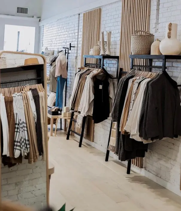 Retail clothing store interior with white brick walls, wooden clothing racks displaying neutral-toned garments, and woven baskets and pottery on upper shelves