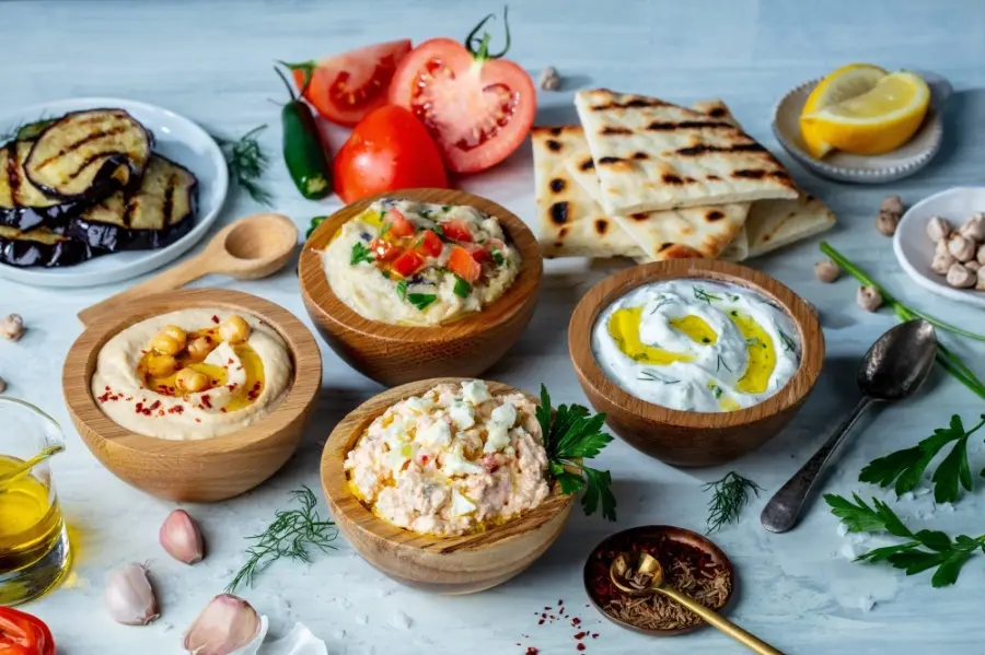 Hummus, tzatziki, and whipped feta dips in wooden bowls surrounded by grilled pita, fresh tomatoes, olives, and herbs on a blue surface