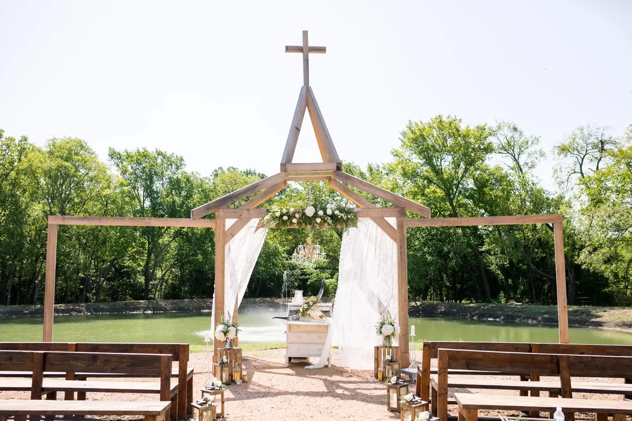 Wooden wedding chapel with cross on top, white fabric draping, wooden benches on either side, surrounded by green trees