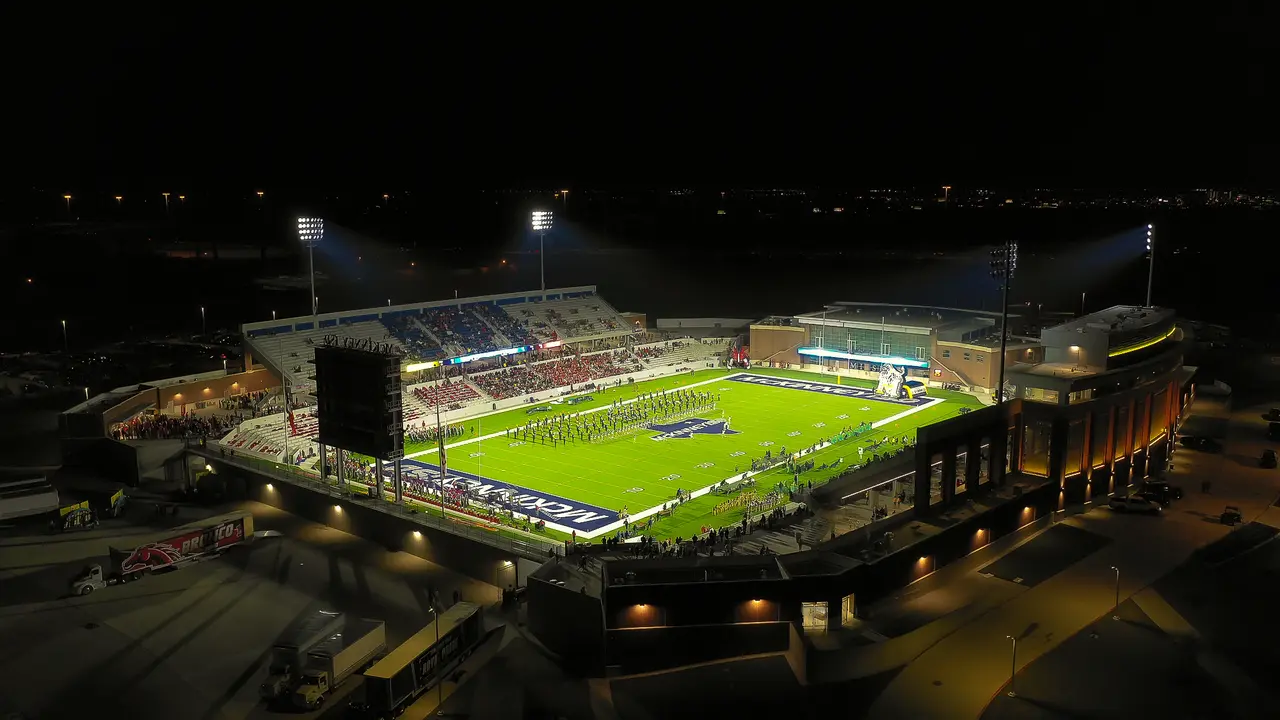 Night aerial view of illuminated football stadium with field, seating, and surrounding buildings
