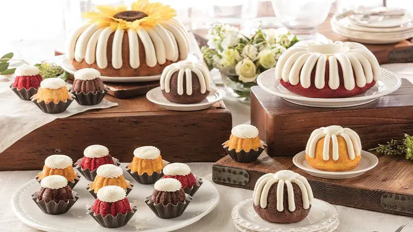 Assorted bundt cakes in various sizes with white glaze and filling, displayed on wooden blocks and white plates with fresh flowers