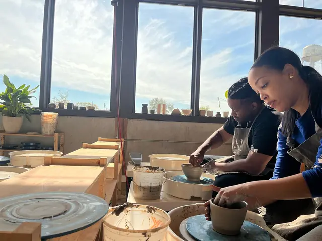 Two people working with clay at a pottery wheel in a bright studio with large windows overlooking a city skyline