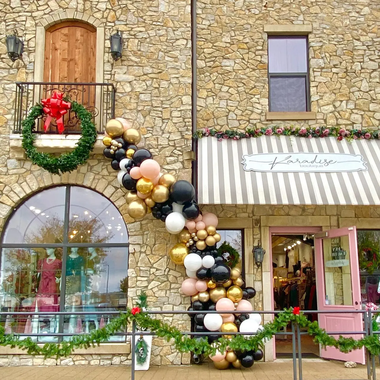 Stone storefront with decorated entrance featuring wreath with red bow, striped awning, and balloon garland in gold, black and white cascading down the facade