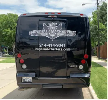 Black charter bus parked in residential driveway with company logo and contact information on rear panel, trees and grass visible in background