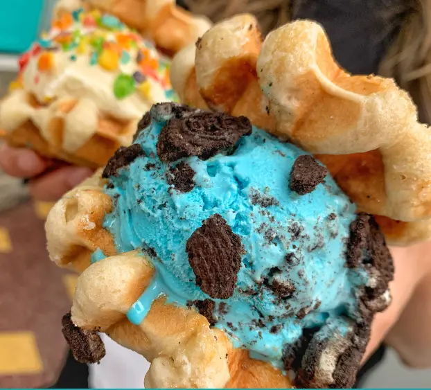 Bright turquoise ice cream with chocolate cookie pieces sandwiched between two waffle cones, held in someone's hand