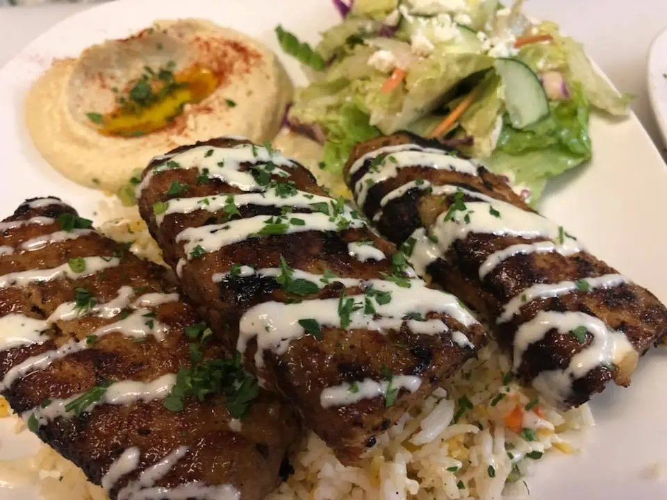 Grilled meat skewers with white drizzle and fresh herbs, served with hummus and green salad