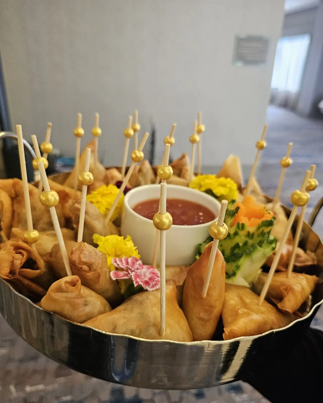 Fried appetizers on wooden picks arranged in a metal bowl around a central sauce cup, garnished with flowers and herbs