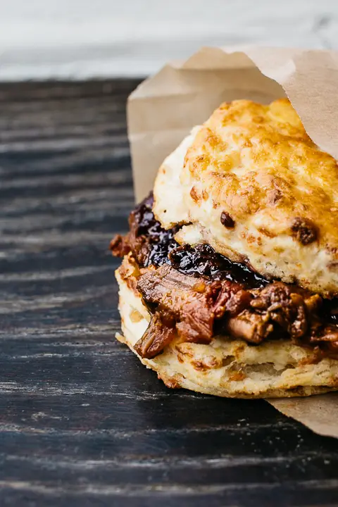 Toasted biscuit sandwich filled with braised meat and topped with melted cheese, wrapped in paper on dark wood surface