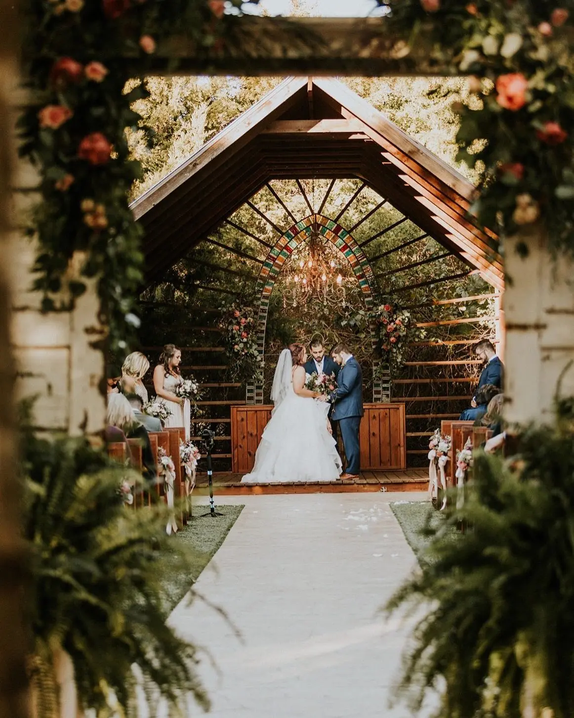 Bride and groom exchanging vows beneath wooden arched pergola adorned with climbing vines and orange flowers, guests seated on either side