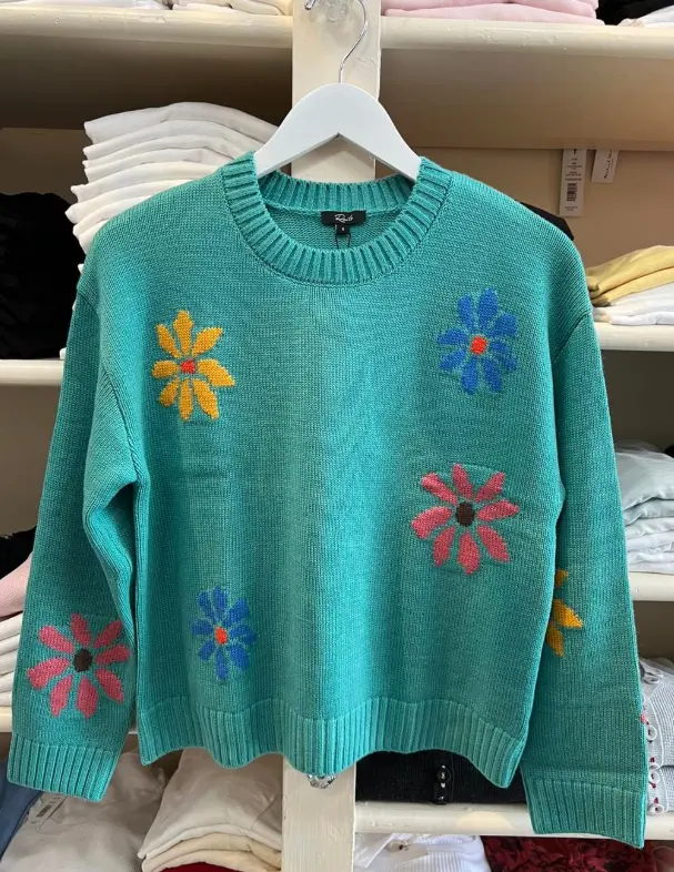 Turquoise knit sweater with colorful embroidered flower motifs displayed on white hanger in retail shop with folded linens on shelves behind