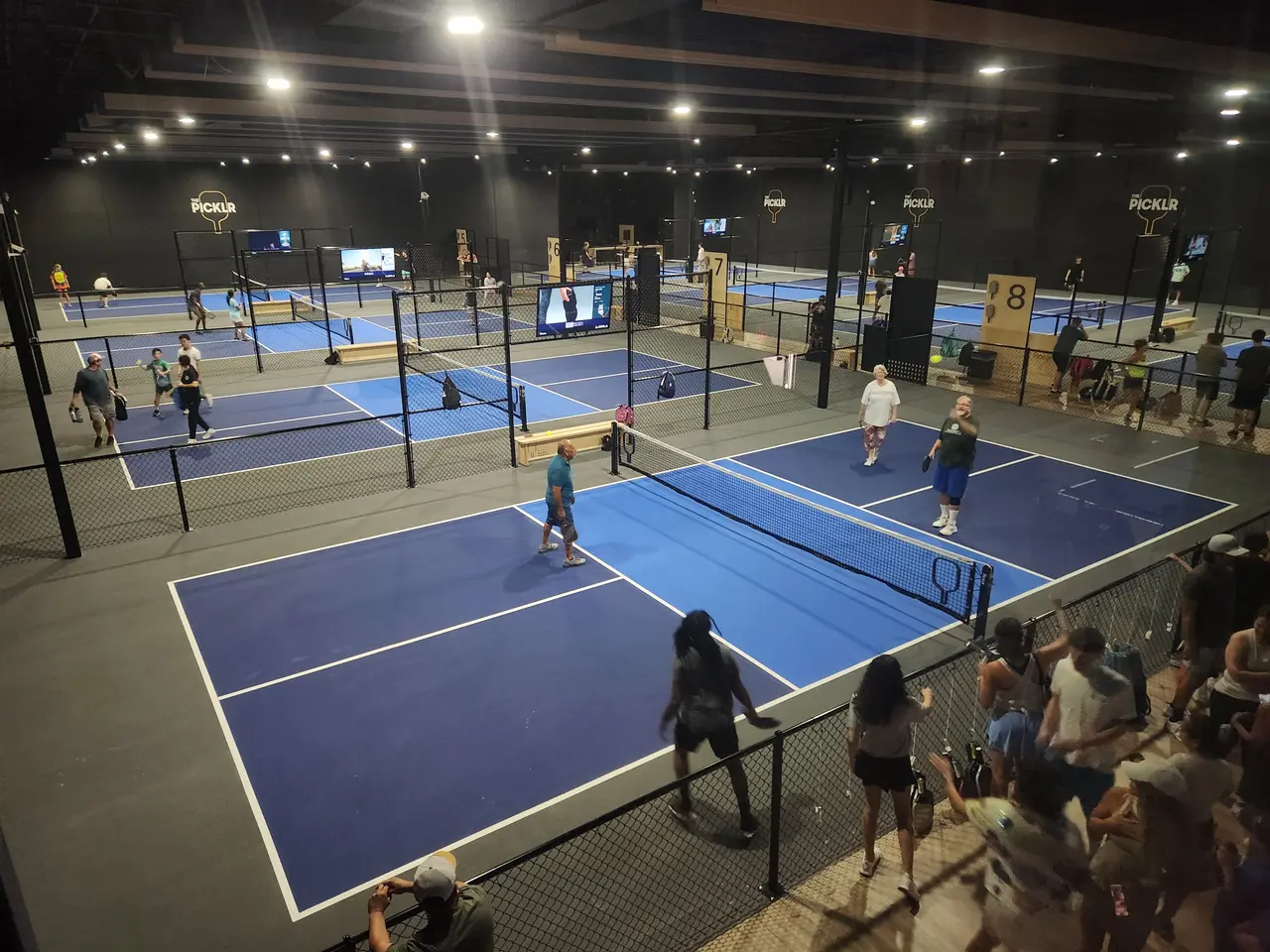 Indoor pickleball facility with multiple blue and yellow courts, players actively engaged in games, and spectators watching from the sidelines