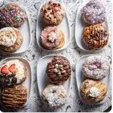 Nine glazed and decorated doughnuts arranged in three rows, each in a paper wrapper, displaying various toppings and finishes
