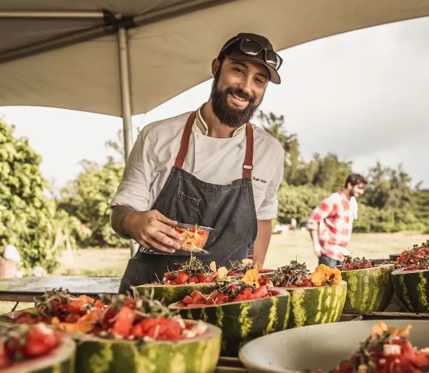 Man in apron and cap arranging fresh strawberries at outdoor catering station under tent, with helpers and green fields visible behind