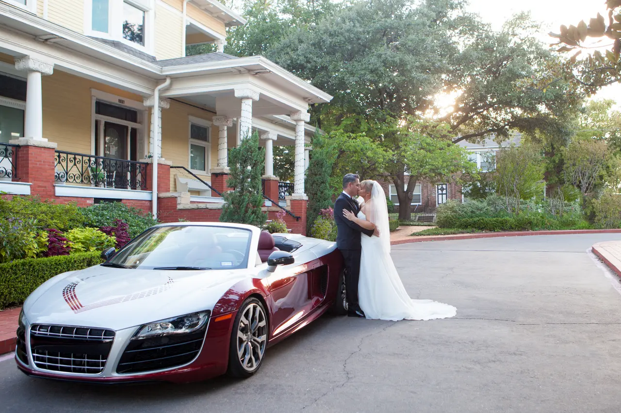 Bride and groom posing with red sports car in front of historic mansion with columned porch and manicured gardens