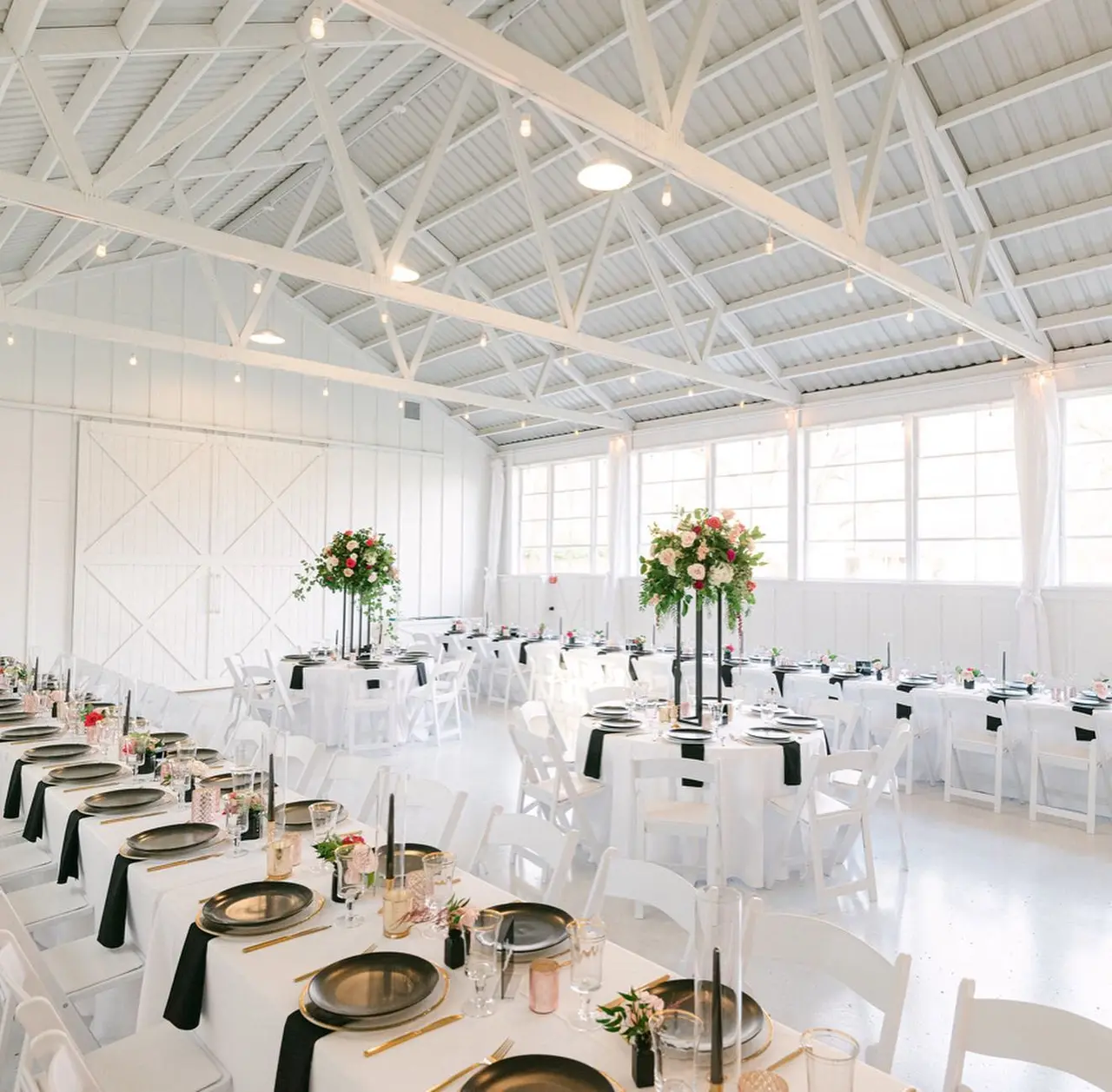Bright white barn interior with high exposed beam ceiling, long banquet tables set with black napkins and gold plates, tall floral centerpieces, and white