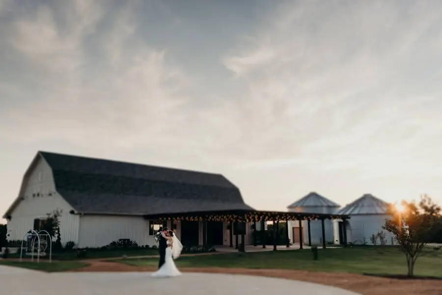 Bride and groom standing in front of a white barn with covered pavilion and grain silos at sunset