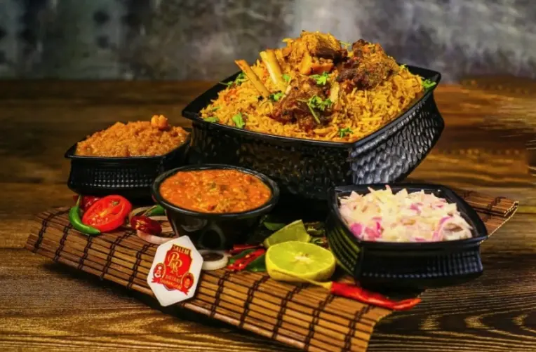 Biryani and curries in black bowls with fresh tomatoes, lime, and chili peppers on wooden surface