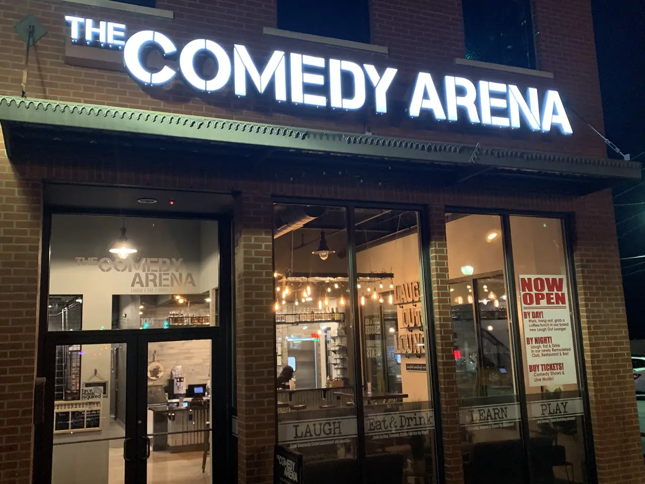 Illuminated storefront of comedy venue at night with brick facade and glass doors, warm interior lighting visible through windows