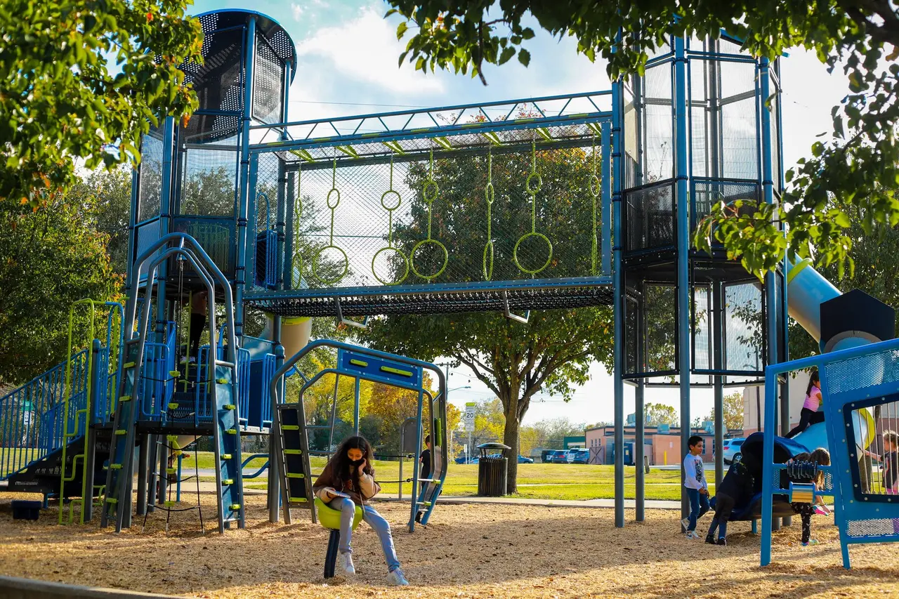 Children playing on blue playground equipment with climbing structures and slides in a park setting surrounded by trees