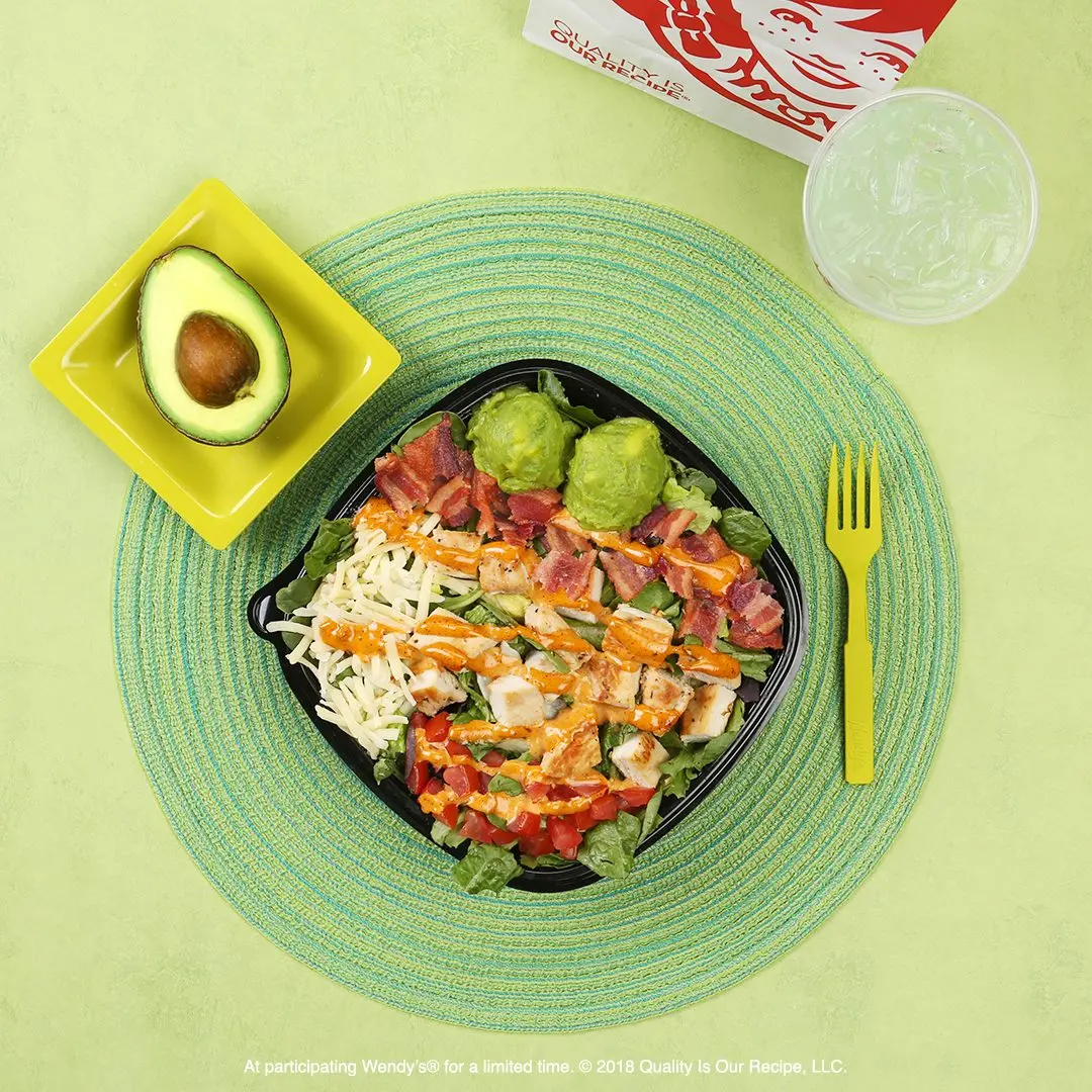 Salad bowl with shredded chicken, lettuce, tomatoes, and carrots on green placemat, with avocado half and water glass nearby