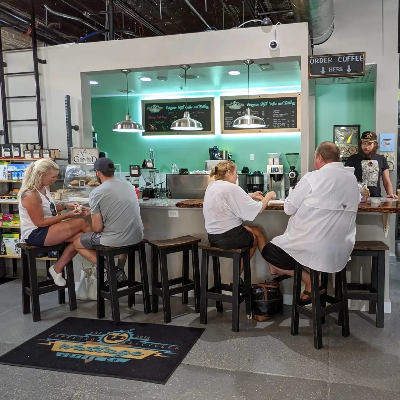 Customers seated at a counter with black stools facing a bright green wall with espresso machine and menu boards, pendant lights overhead