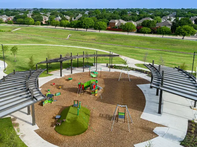 Aerial view of circular playground with colorful climbing structures, shade structures, and seating surrounded by open grass fields and suburban homes