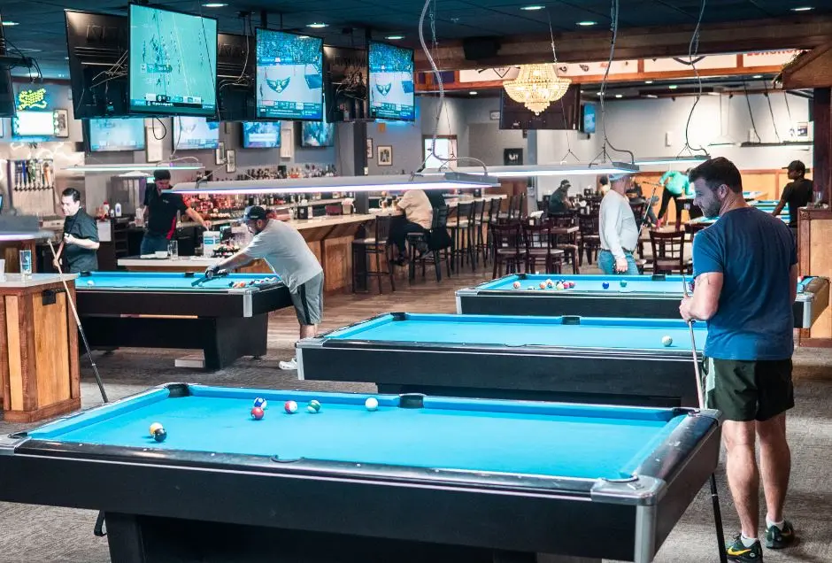 Interior of sports bar with multiple pool tables, players, bar counter, and TV screens mounted on walls