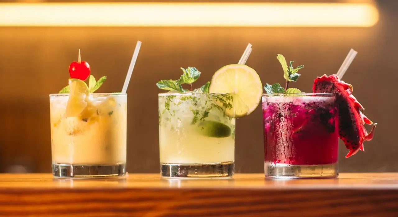 Three colorful cocktails with straws and garnishes on a wooden bar counter against warm lighting