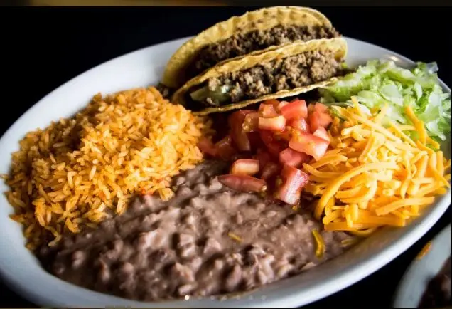 Plated Mexican food with two tacos, rice, beans, shredded cheese, lettuce, and diced tomatoes